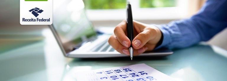 Mão segurando uma caneta sobre um calendário e um notebook ao fundo